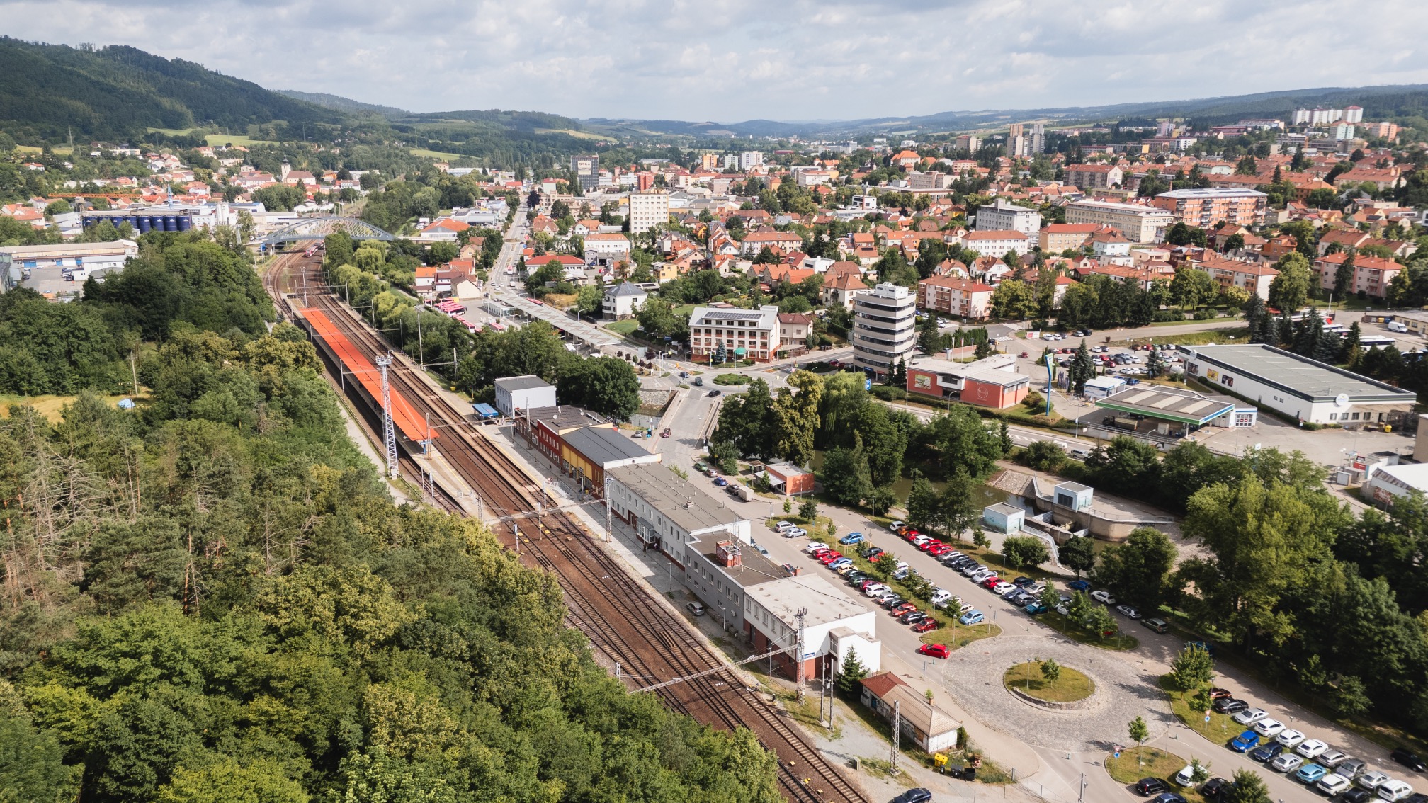 Aerial view of a small town with railroad tracks running alongside buildings and green wooded areas under a partly cloudy sky.
