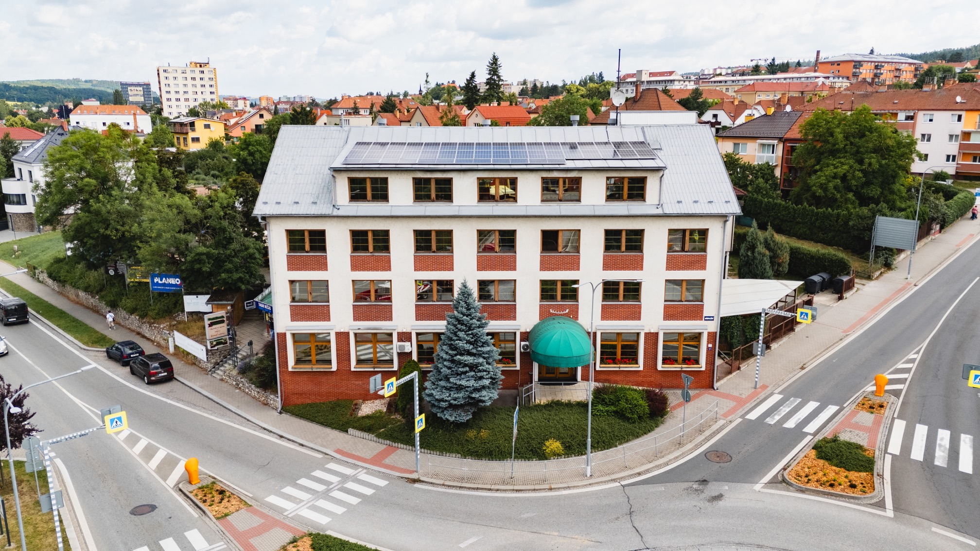 Three-story building with solar panels on the roof, a green awning entrance, and a blue spruce tree in front, located at an intersection with crosswalks.