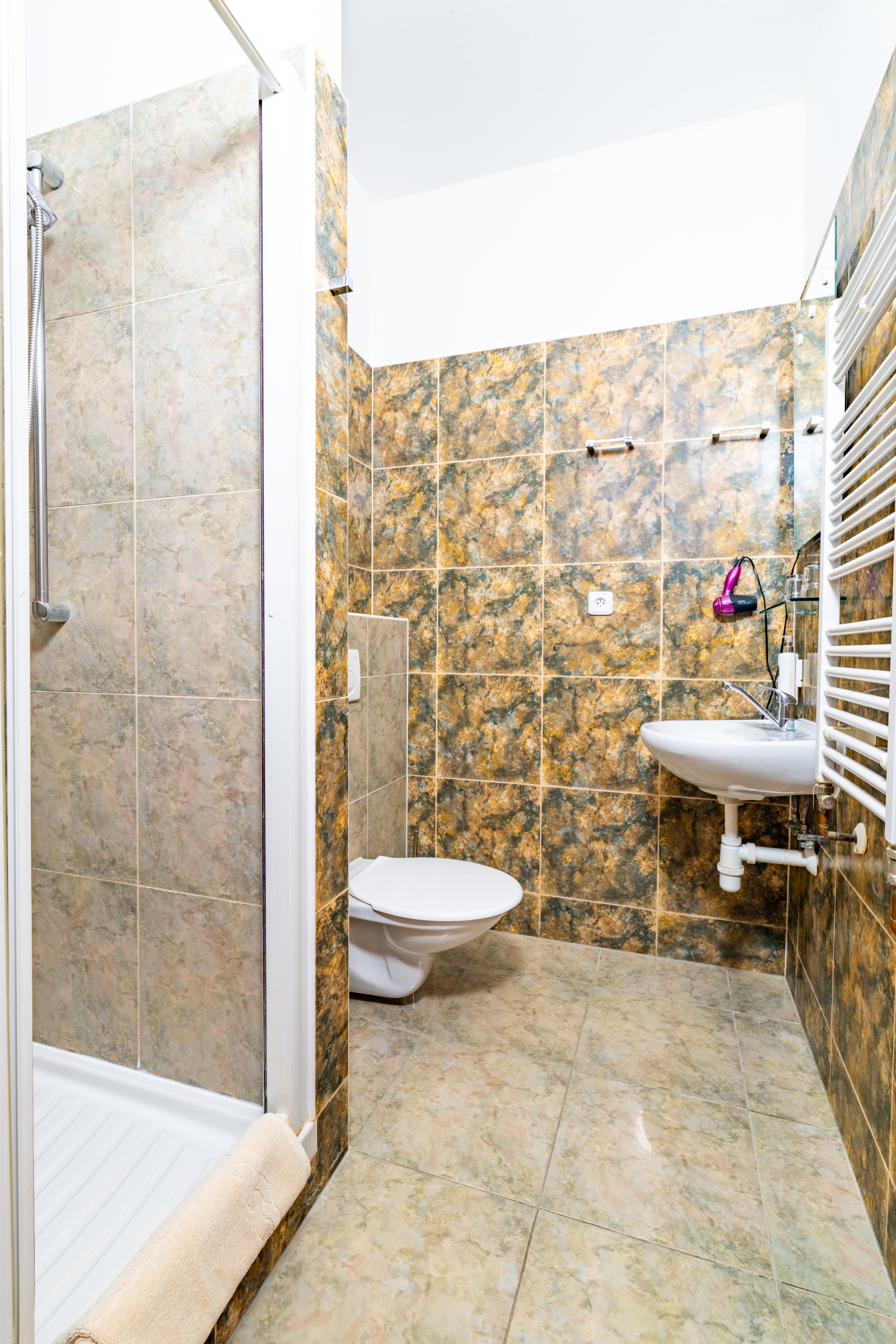 Small bathroom with a wall-mounted toilet, a sink, a towel radiator, and a shower stall with beige and brown marble tiles.