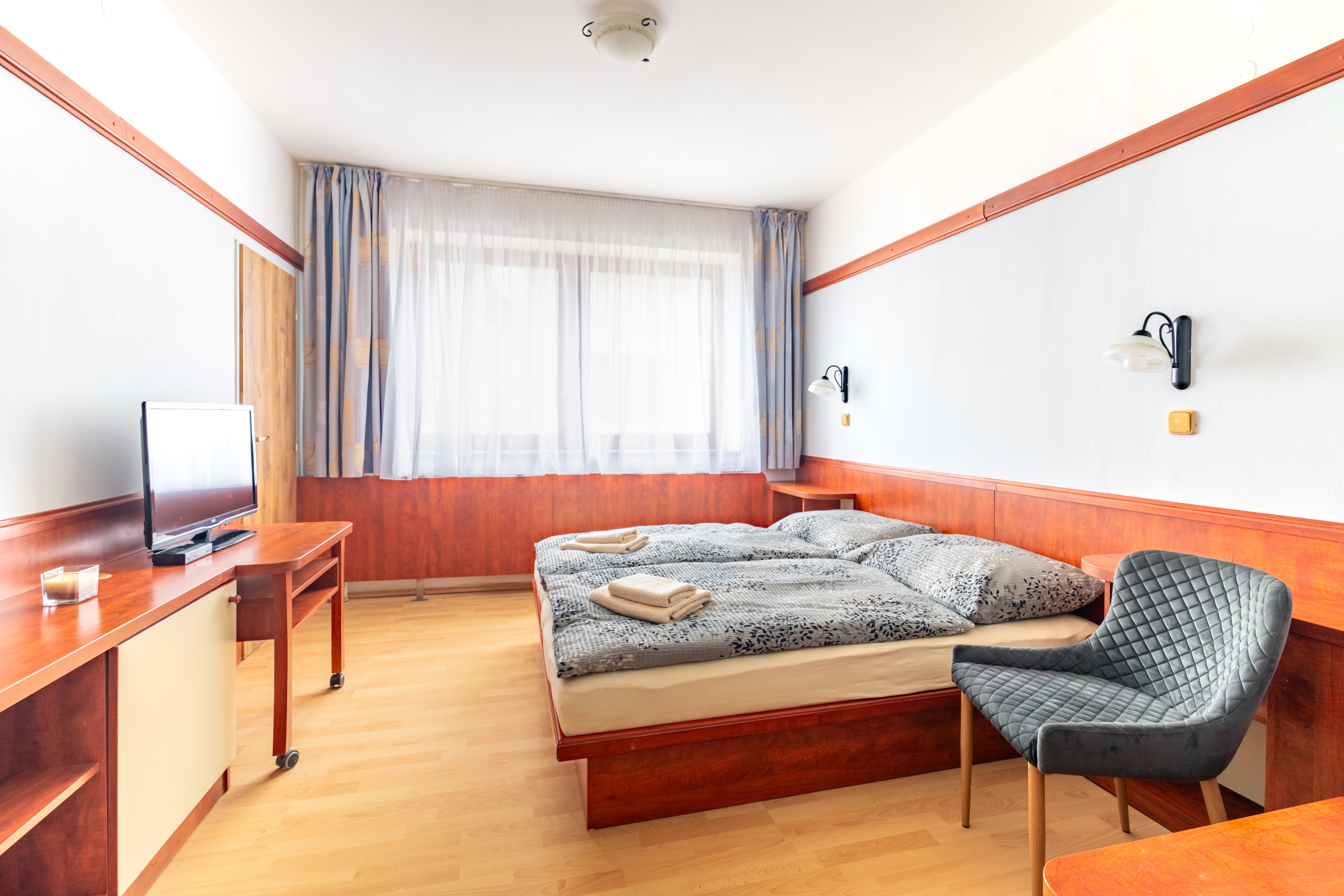 Bright hotel room with a double bed, patterned gray bedding, folded towels, a gray quilted chair, and a TV on a wooden desk.