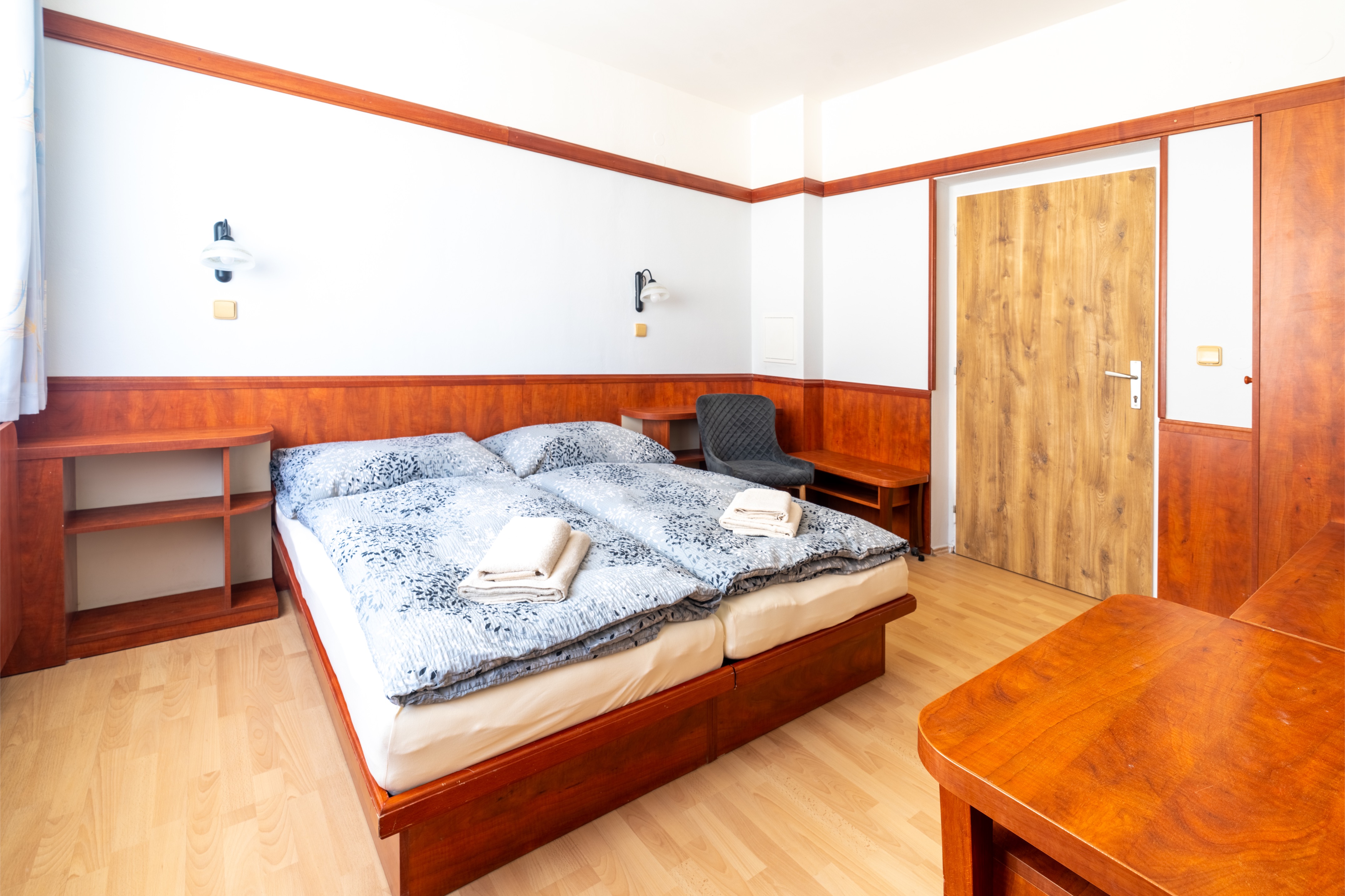 Minimalist bedroom with twin beds pushed together, light blue patterned bedding, wooden furniture, and a dark gray chair.