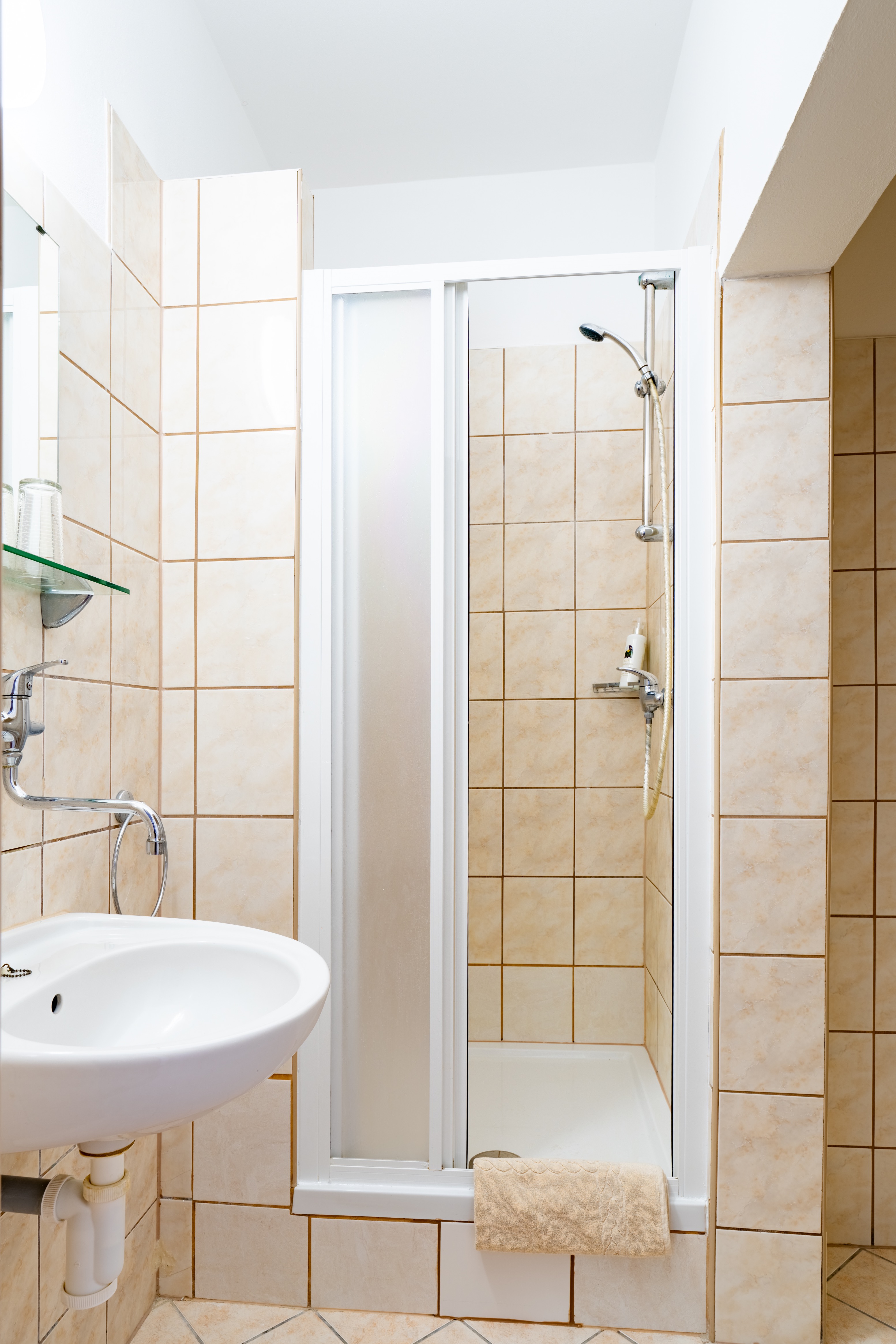 Compact bathroom with a white sink, wall-mounted faucet, and a tiled shower with a sliding glass door and beige towel.