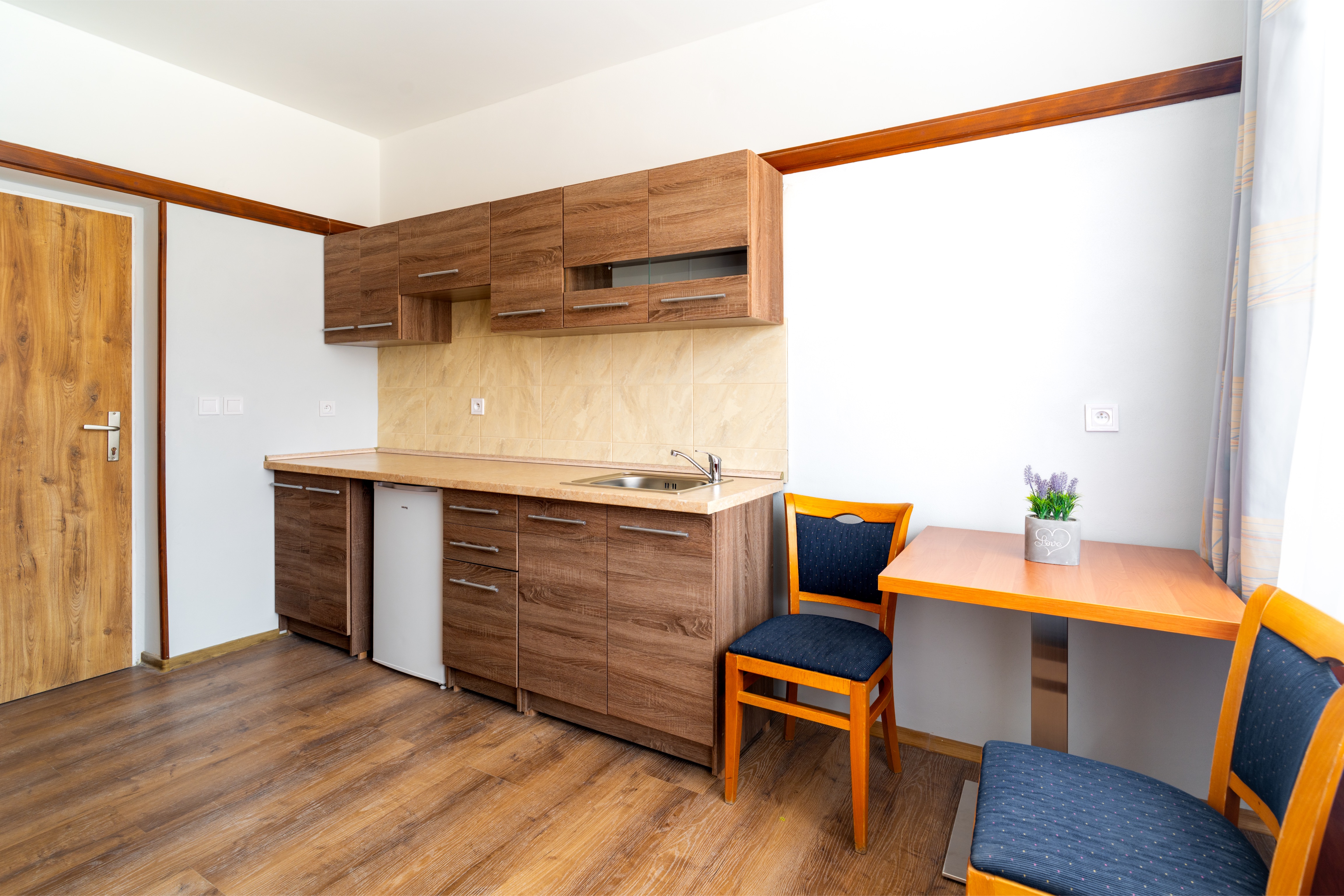 Small kitchen with wooden cabinets, a countertop with a sink, mini fridge, and a dining table with two blue cushioned wooden chairs and a small plant.