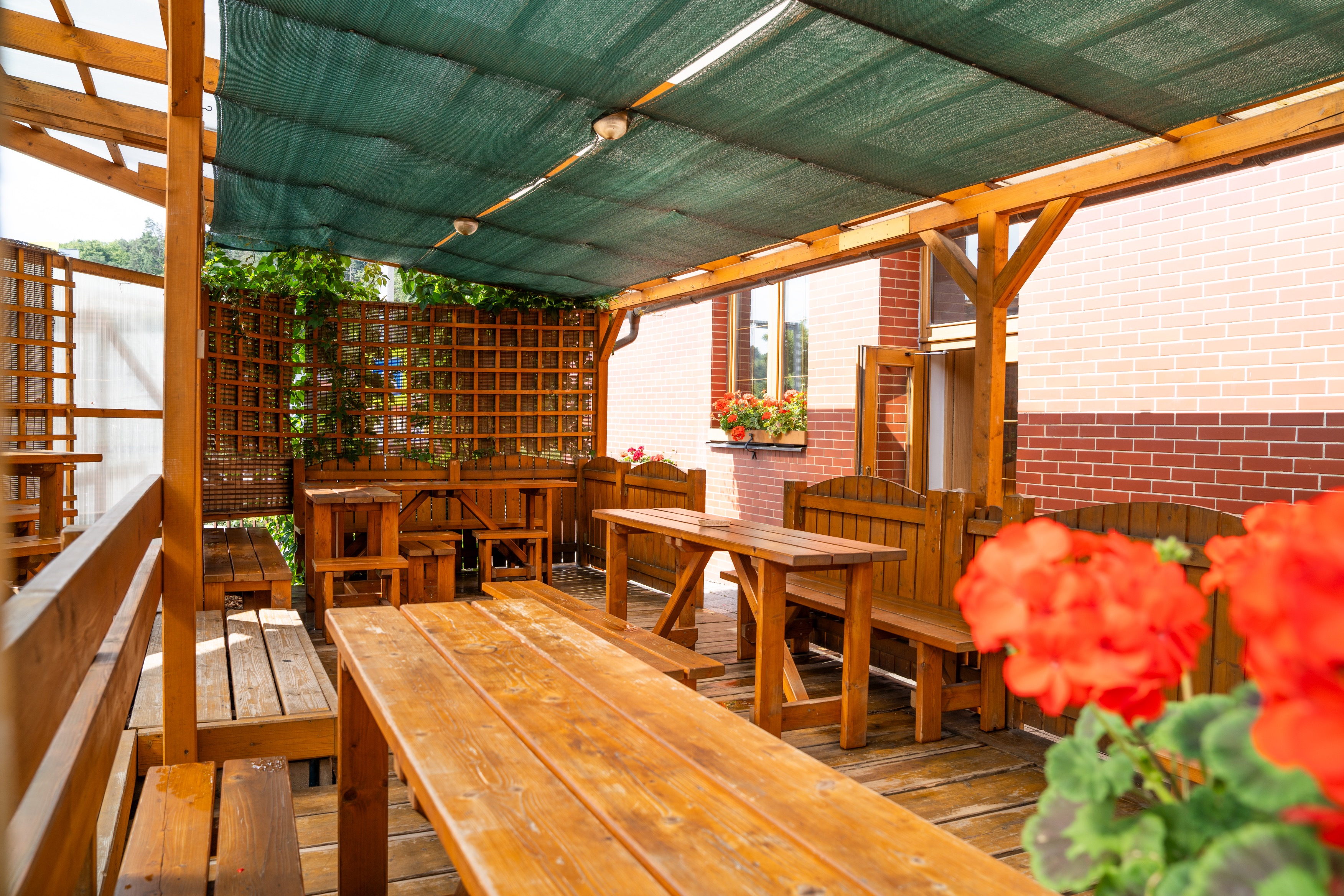 Outdoor wooden patio with tables and benches under green fabric shade and red flowers in the foreground and window boxes.