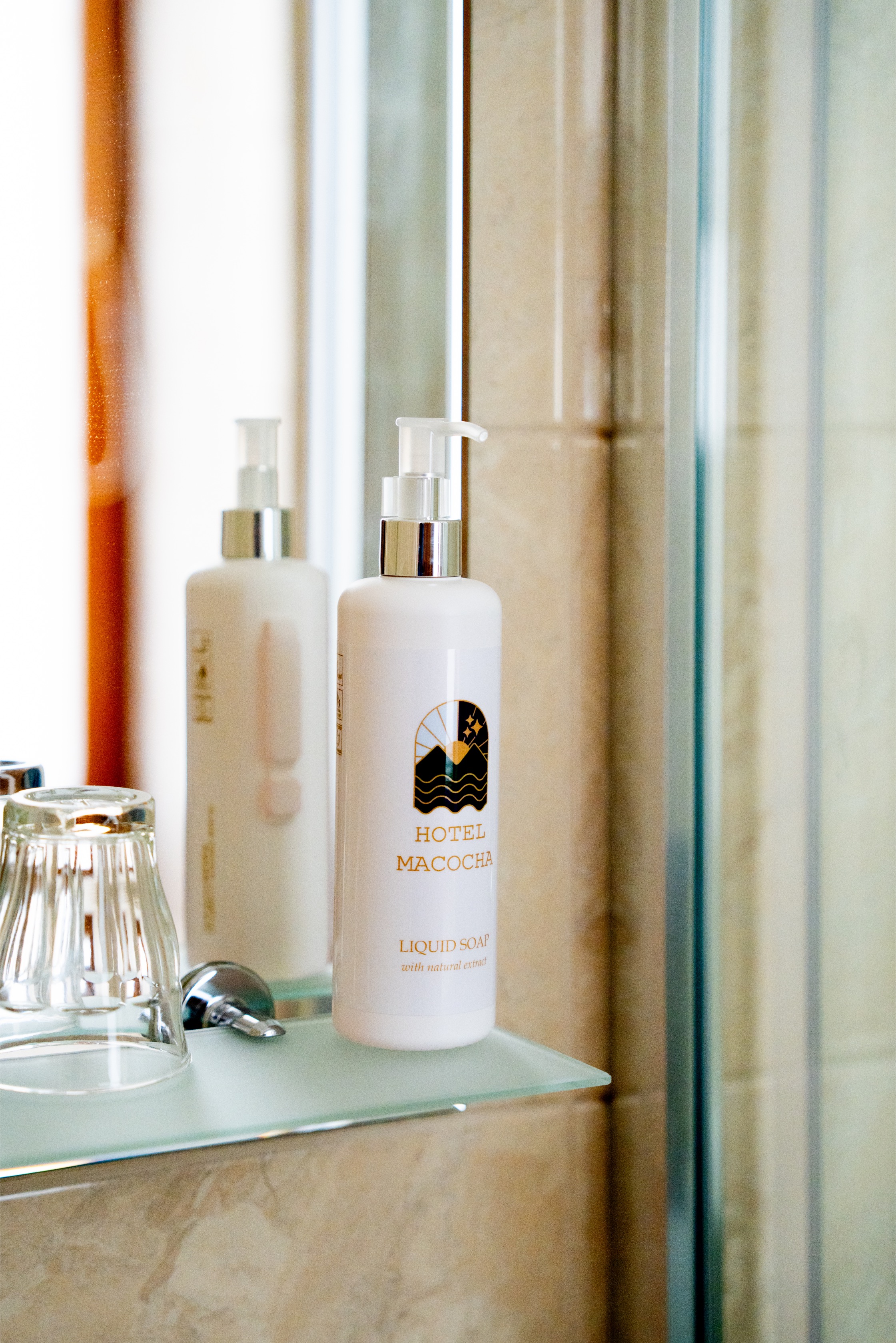 A white pump bottle of Hotel Macocha liquid soap on a glass bathroom shelf next to an inverted glass and reflected in a mirror.