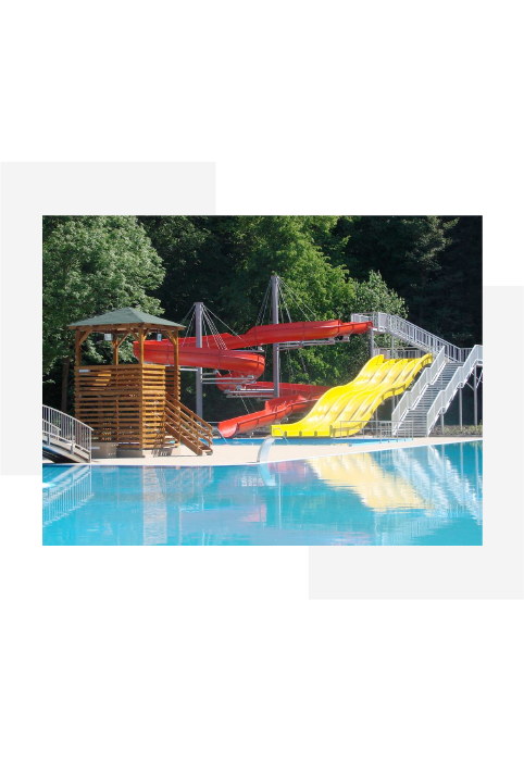 Outdoor water park with red spiral and yellow straight water slides next to a clear swimming pool surrounded by trees.
