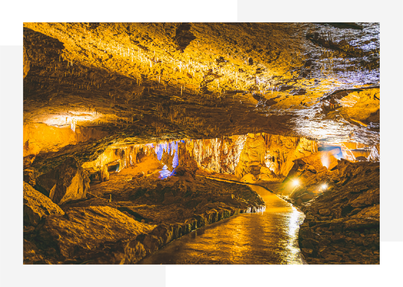 Illuminated cave interior with a paved pathway, stalactites hanging from the ceiling, and stalagmites rising from the floor.