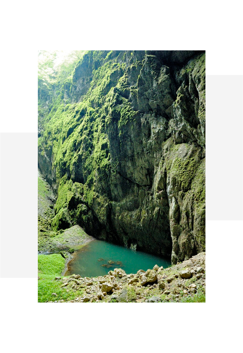 A deep canyon with steep rocky walls covered in green moss surrounding a small turquoise pool of water at the base.