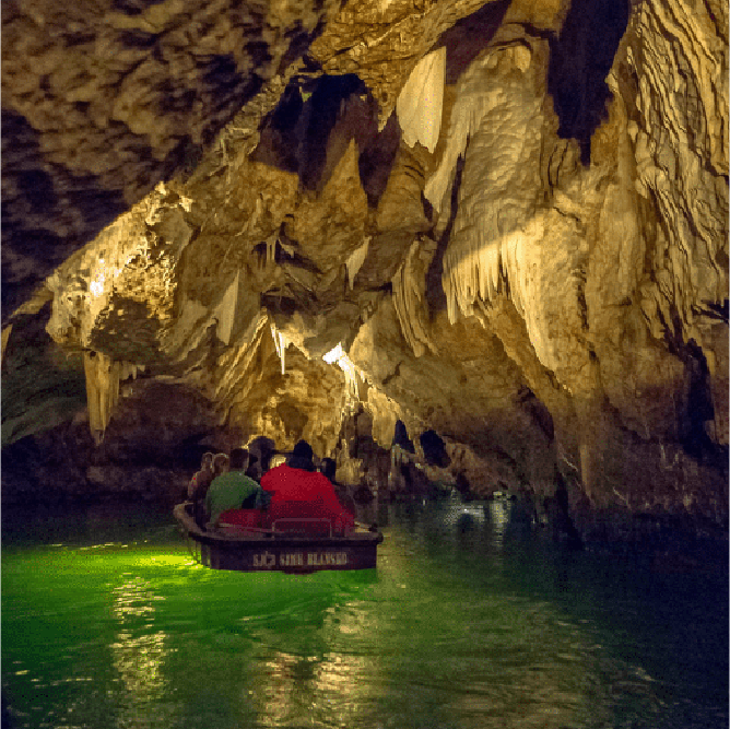Group of people in a small boat navigating through an illuminated cave with stalactites above and water below.