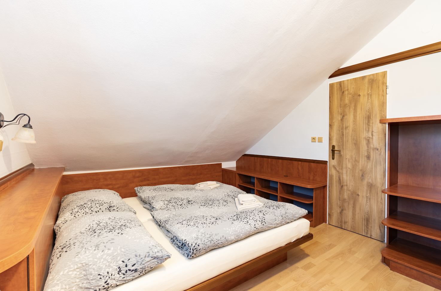 Cozy attic bedroom with wooden furniture, a double bed with gray floral bedding, and folded white towels on top.