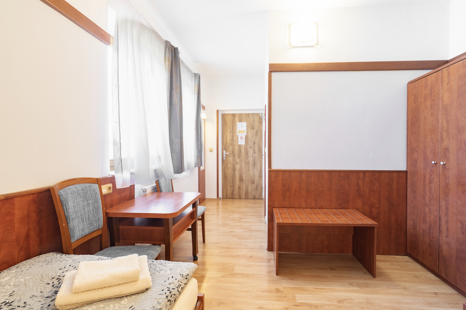 Minimalist hotel room with wooden furniture, a single bed with folded towels, two chairs, a table, a wooden bench, and a wardrobe.