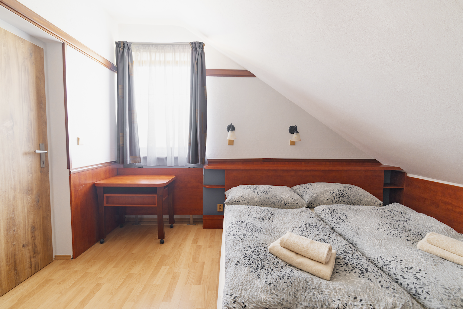 Small attic bedroom with two single beds pushed together, gray patterned bedding, folded beige towels, wooden desk, window with gray curtains, and slanted ceiling.