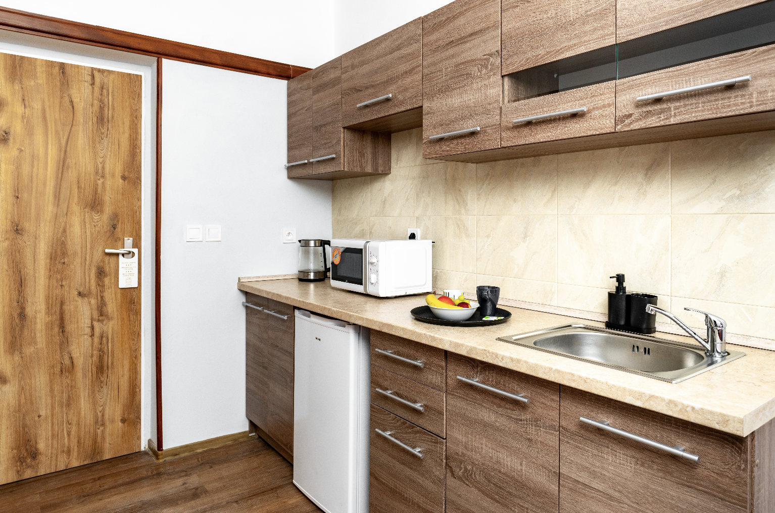 Compact kitchen with wooden cabinets, a countertop holding a microwave, electric kettle, fruit bowl, cup, and a stainless steel sink with soap dispensers.