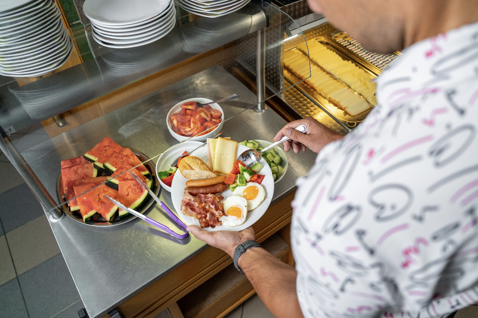 Person holding a plate with sunny-side-up eggs, bacon, sausages, bread, cheese slices, and a vegetable salad near a buffet with watermelon slices and bowls of salad.