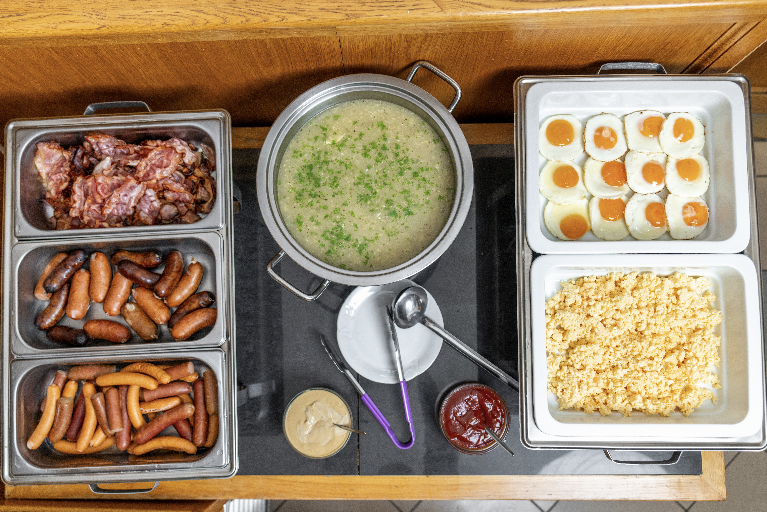Buffet with cooked bacon, sausages, scrambled eggs, fried eggs, a pot of soup garnished with herbs, mustard, and ketchup.