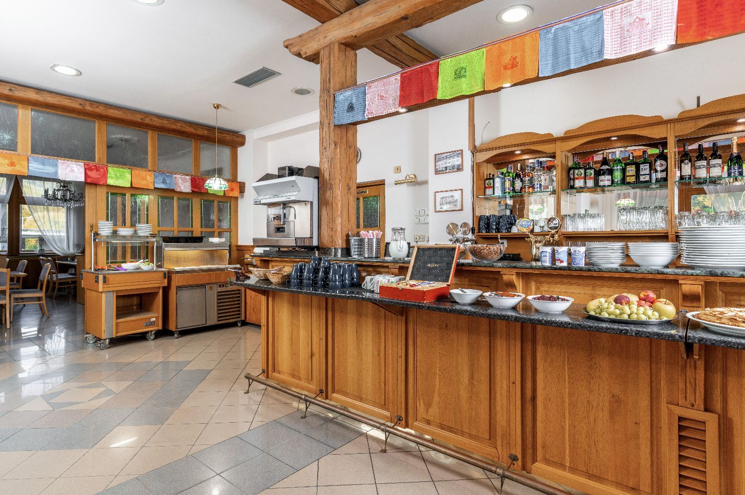 Interior of a wooden counter buffet area with bowls of fruits and cereals, coffee mugs, and bottles on shelves in a bright dining room.