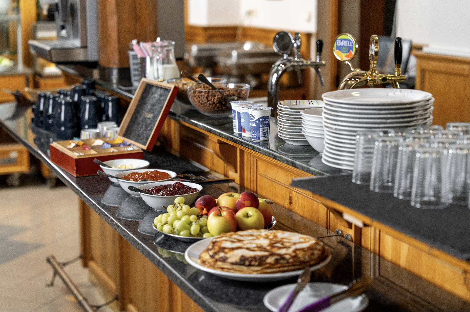 Breakfast buffet with pancakes, apples, grapes, bowls of jam, yogurt cups, plates, glasses, and coffee mugs on a counter.